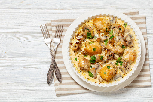 chicken thighs in a creamy mushroom garlic sauce with herbs and parmesan cheese in white baking dish on white wooden table with forks, horizontal view from above, flat lay, free space