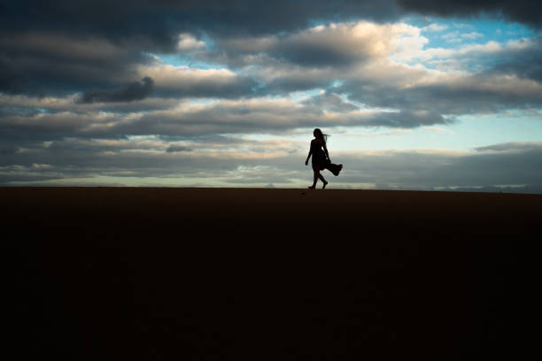 A woman is walking on a sandy beach at dusk. The sky is cloudy and the sun is setting