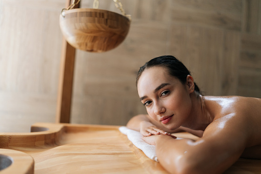 Portrait of tranquil young woman getting massage lying relaxing on massage table, looking at camera. Happy carefree lady enjoying leisure in wellness salon. Concept of spa health and self-care.