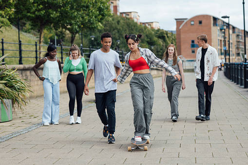 Wide shot of a group of teenage friends walking along the Quayside in Newcastle Upon Tyne. The girl in the front is riding a skateboard while holding her friend's hand for support. The girl in the back is on the phone.\n\nVideos are available similar to this scenario.