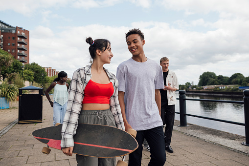 Medium shot of two teenage friends walking side by side while smiling and looking at each other. The girl is carrying a skateboard under her arm. The rest of the group is walking behind them.\n\nVideos are available similar to this scenario.