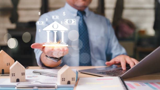 istock Virtual banking manager is holding a bank symbol in his hand while sitting at a desk with a laptop. Concept of financial stability and success 2148262342