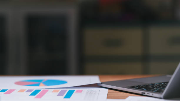 istock A laptop sits on a table with a stack of colorful graphs. The graphs are arranged in a pie chart and a bar chart, and they appear to be related to some kind of data analysis or financial report 2148262343