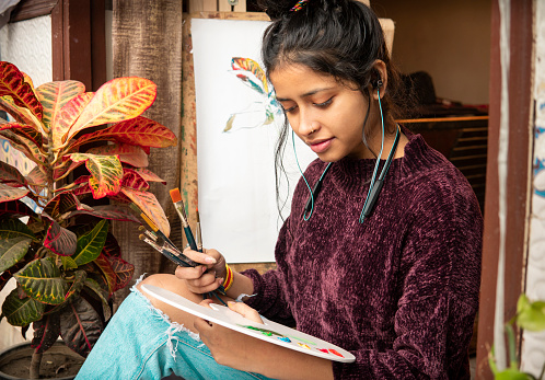 Happy Asian, Indian beautiful female artist doing painting on canvas with water color and paintbrush and listening music through earphones in her leisure time at home.
