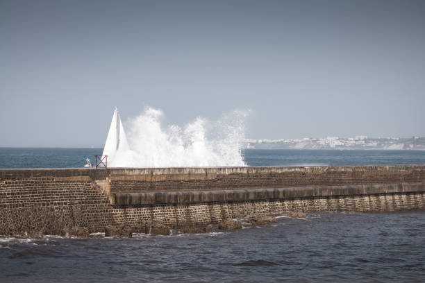 white boat sails peeking out behind a stone dike with a splash of wave