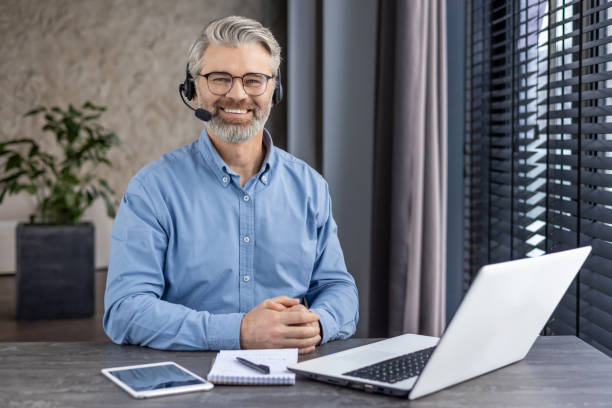 Portrait of mature successful gray haired man inside office at workplace inside office, businessman with headset phone and laptop for video call smiling and looking at camera