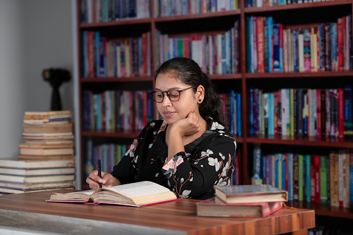 Young female reader in the library, exploring, reading various books, taking notes, and concentrating. The young reader with spectacles, hair nicely clipped behind, wearing a black shirt with a white floral pattern, exploring various books in the library. Sitting on the chair with a book placed on the brown top table, reading with concentration, resting her chin on her left palm, and marking on the book with a pen. A few books are kept in the foreground, and a stack of old books is placed in the long corner in the background with the bookshelf.