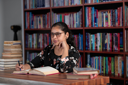 Young female reader in the library, exploring, reading various books, taking notes, and concentrating. The young reader with spectacles, hair nicely clipped behind, wearing a black shirt with a white floral pattern, exploring various books in the library. Sitting on the chair with a book placed on the brown top table, reading with concentration, resting her chin on her left hand, and pointing on the book with a pen, looking straight towards the light. A few books are kept in the foreground, and a stack of old books is placed in the long corner in the background with the bookshelf.