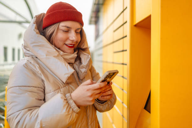 istock Happy smiling woman client with box at outdoor automated parcel machine choosing operation on touch screen by smart phone 2148264946