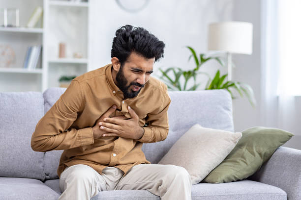 istock Frightened arab male keeping crossed hands on chest while squinting face in grimace of ache in apartment interior. Young man experiencing acute pain and risk of heart attack symptoms on sofa 2148266806