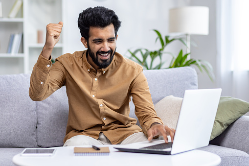 Excited hispanic freelancer raising hand with clenched fist while typing on laptop keyboard at home office. Rejoiced man showing winner gesture while answering mail with profitable project deal.