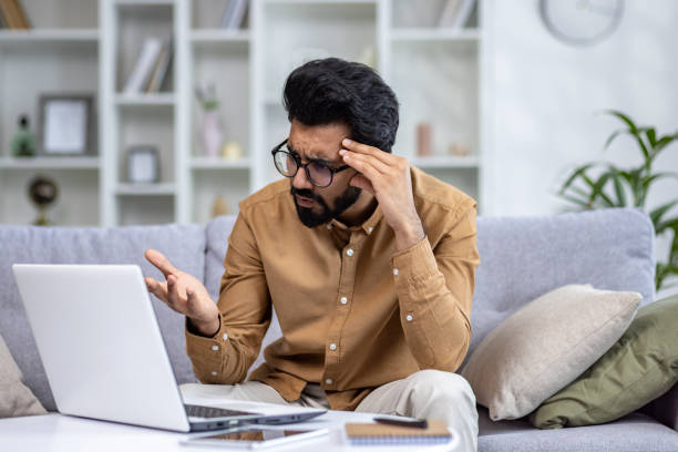 istock Worried arab male in eyeglasses looking at portable computer screen with confusion while sitting at pillowed sofa. Stressed technical student struggling with learning information from remote class 2148266826