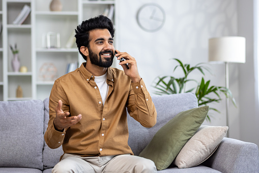 Smiling islamic man sitting on grey pillowed sofa and talking by mobile phone while looking aside. Satisfying male relaxing at home and having pleasing distant conversation by wireless device.