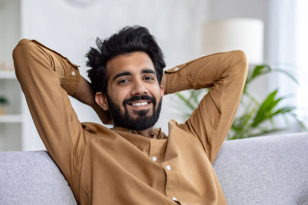 istock Positive muslim guy leaning on melange couch with hands behind head and smiling at camera on blurred background. Handsome man spending free time at home and enjoying break at renovated furniture 2148266839