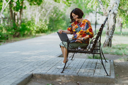 Female professional enjoys remote work on a calm sunny day in a lush green park. Nature meets technology with a fashionable woman using her laptop outdoors on a sunny day