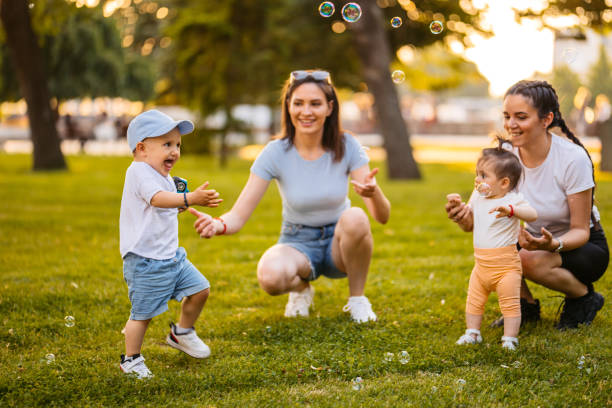 Two Female Friends Blowing Soap Bubbles With Their Babies In The Park