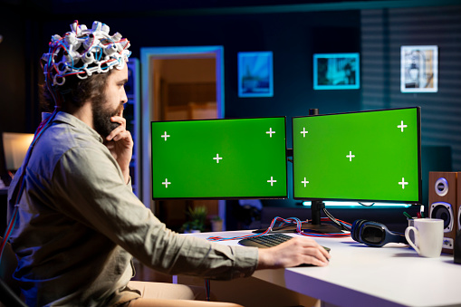 Man with EEG headset programs brain transfer into multimonitor setup green screen computer, merging with artificial intelligence. IT expert uses neuroscience and mockup desktop PC to gain digital soul