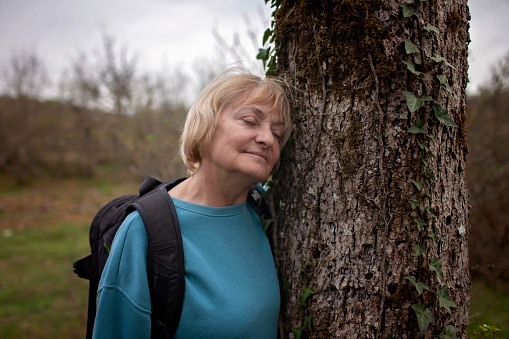 A senior woman with a serene expression rests against an old tree trunk, eyes closed, immersed in the tranquility of nature during a hiking break.
