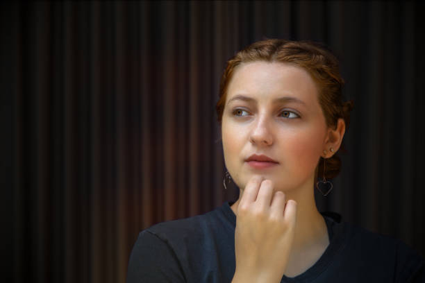 A pensive woman contemplates with hand on chin, her expression thoughtful and serious, against a dark background.