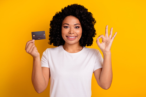 Photo of optimistic woman with chevelure wear white t-shirt recommend you credit card showing okey isolated on yellow color background.