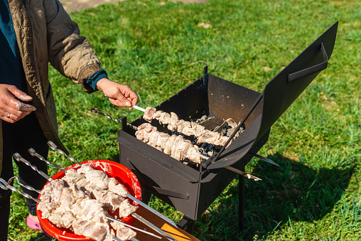 Woman strands raw pieces of marinatedo pork meat on long skewers frying shish on the grill.Nice summer day.