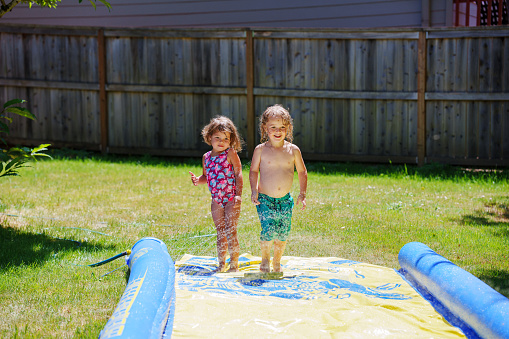 A three year old Eurasian girl and her boy cousin who is the same age stand together while playing on a slip n slide in the back yard on a hot summer day in Oregon.