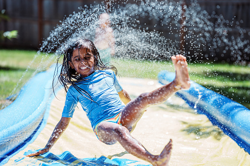 An adorable young Indian girl laughs laughs while playing on a slip n slide in the backyard of her home on a hot and sunny summer day.