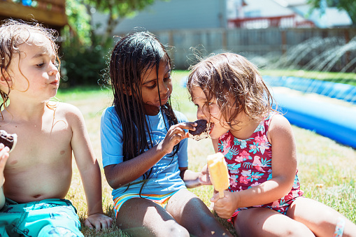 An Kindergarten age Indian girl shares a bite of her ice cream sandwich with her friend while sitting outside in the grass on a hot summer afternoon. The multiracial group of friends are wearing wet swim suits because they have been playin in the sprinklers together.