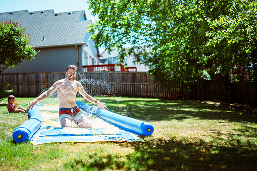 A Caucasian man laughs while sliding down a slip and side in his backyard on a summer day, while playing outside with his Eurasian toddler daughter who is sitting in the grass and watching him.