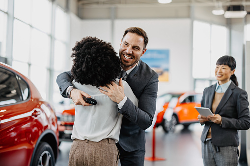 In the car showroom, a multiracial couple shares an embrace, their closeness reflecting the unity of their dreams and the excitement of embarking on new journeys together, surrounded by the possibilities each car represents