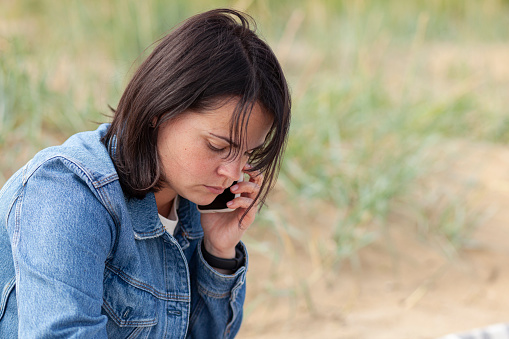 A beautiful dark-haired girl in a denim jacket speaks sadly on the phone on the beach near the sea