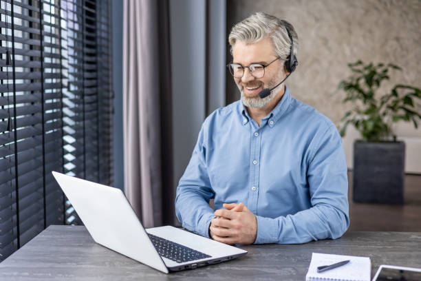 istock Satisfied senior entrepreneur in headset with microphone looking at portable computer screen with hands folded on desk. Pleased boss listening to reports of employees with improved results at home 2148280982