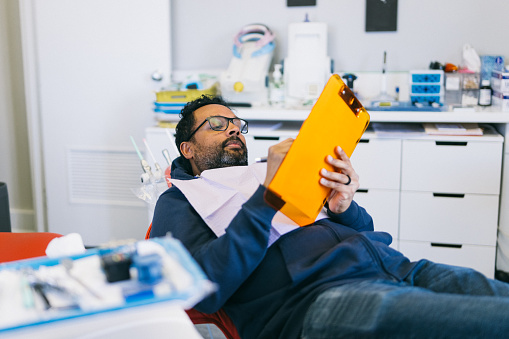 A male patient of Indian descent lays on a dentist chair, filling out paperwork on a clipboard while awaiting his dental appointment.