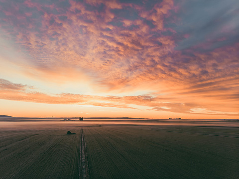 A beautiful sunset over a field with a lone tractor in the foreground. The sky is filled with clouds and the sun is setting, creating a serene and peaceful atmosphere