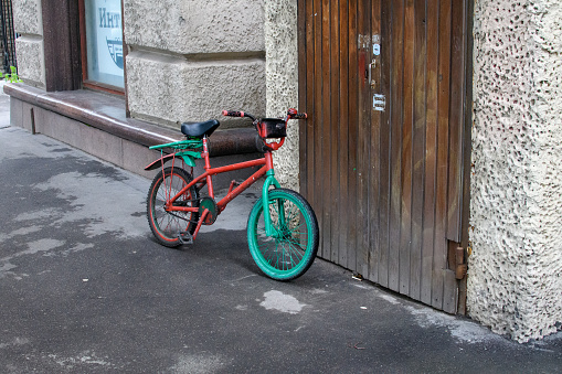 Moscow, Russia - 29 Aug 2017: A vibrant red and green bicycle stands alone, leaning against a textured wall, embodying the quiet stillness of a city street. This image captures a colorful bicycle abandoned beside a locked wooden door and textured wall on an empty street; its solitude and vibrancy contrasting the silent urban surroundings.