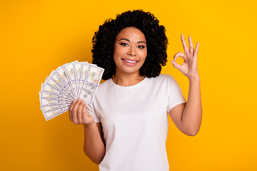 Photo of good mood woman with chevelure wear white t-shirt holding dollars showing okey good work isolated on yellow color background.
