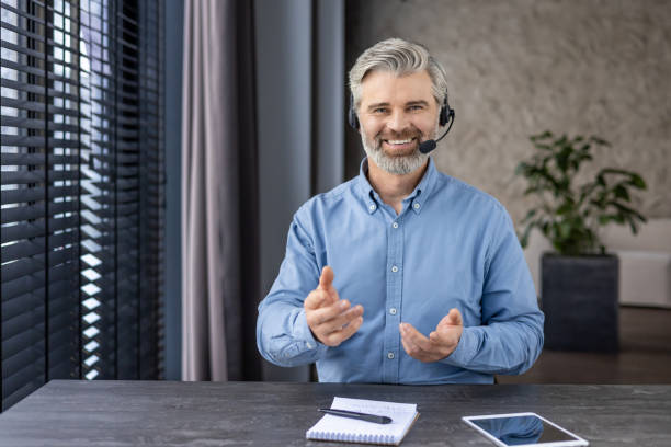 istock Webcam view, mature successful gray haired boss looking at camera with headset phone inside office at workplace, man smiling talking on online meeting, senior businessman in shirt 2148284218