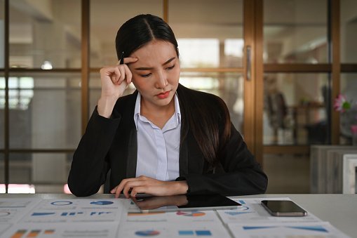 Stressed asian female accountant with papers and charts sitting at her office desk.