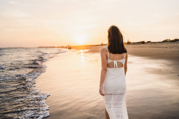 istock beautiful woman walking along the beach shore with sunset light dressed in white 2148286138