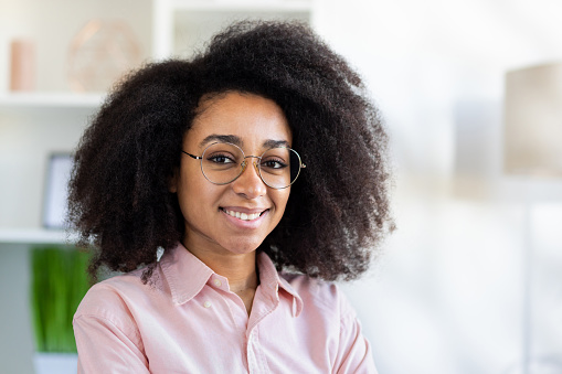 Portrait of beautiful woman with curly hair, African American woman smiling and looking at camera, sitting on sofa happy in living room at home, video call remote conversation.
