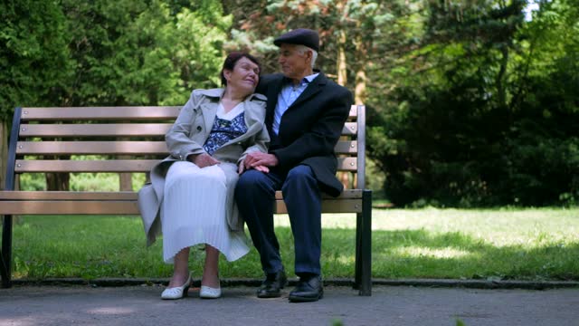 An elderly couple strolling peacefully in the park. A man and woman enjoying a leisurely walk amidst nature. A senior couple cherishing a stroll through the park. An older pair ambling together in a serene park setting. A gentleman and lady leisurely walk