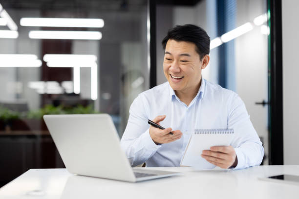 istock Smiling japanese male entepreneur having online video call on modern laptop while holding notebook and pen in hands. Positive man making notes during conversation with interlocutors at office 2148290436