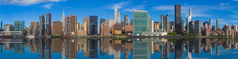 High Resolution Stitched Panorama of New York Skyline with UN Building (Headquarters of the United Nations), Chrysler Building, Empire State Building, Manhattan Upper East Side Residential and Office High-rises, FDR drive, Green Trees and Morning Blue Sky with Clouds All Reflected in Water of East River. Canon EOS 6D (Full Frame censor) DSLR and Canon EF 85mm f/1.8 lens. 4:1 Image Aspect Ratio. This image is downsized to 50MP. The Original image resolution is 111MP or 21031 x 5258 px.