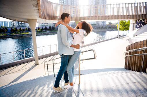 Full body length photo of young family couple spend free time outdoors hugging looking each other bonding laughing at city river pond.
