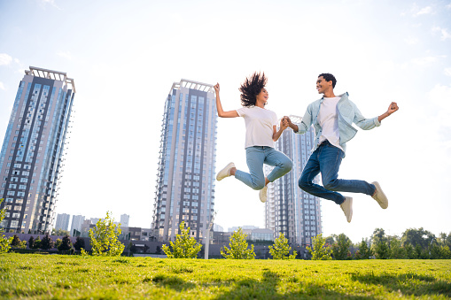 Photo of young happy smiling couple jump active together holding arms american best friends outdoors on modern buildings background.