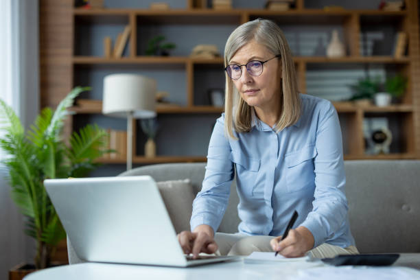 istock Stylish mature woman sitting on couch and using portable computer while writing in notebook at home. Attentive female joining remote classes for advanced training and making remarks from lecturer 2148290809
