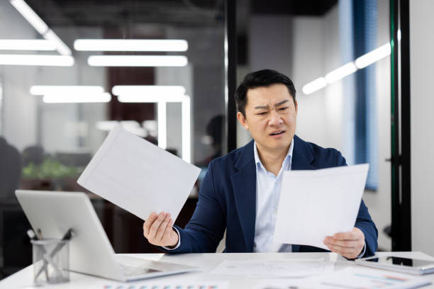 istock Punchy man holding documents while sitting at white desk in office environment. Handsome asian male doing work on background on glassy wall of business center. Concept of technology and people 2148291202