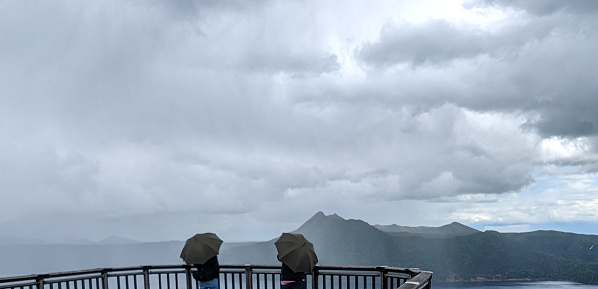 Two senior American travellers take in the panoramic view of Lake Mashu from an observation deck in Akan-Mashu National Park on a misty spring morning. Teshikaga town in Kushiro Subprefecture, Hokkaido Prefecture.