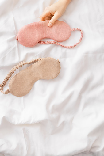 Close up two eye masks for sleep beige and pink color on white crumpled bedclothes, woman hand take one, minimal flat lay. Top view sleeping mask for best sleeper, comfort relax. Rest well concept.