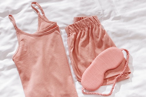 Top view pink pajama and eye sleep mask on white crumpled bedclothes. Cozy pyjamas for comfort rest at night. Flat lay from singlet, shorts, sleeping mask pastel pink color, sleep well concept
